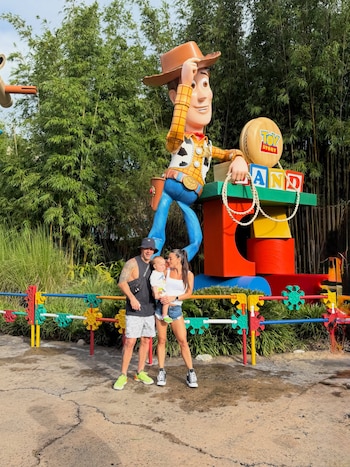 Un hombre, una mujer y un niño pequeño posan frente a una figura gigante del personaje Woody en una zona temática con el letrero Toy Story Land