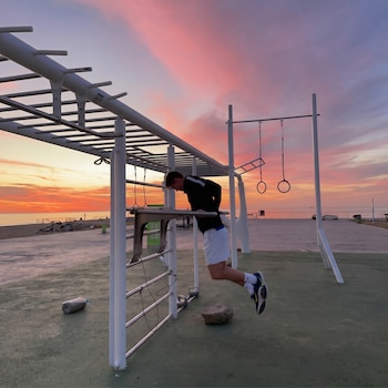 Un hombre realiza dips en barras paralelas de un parque de calistenia, con el mar y un cielo anaranjado y rosado al atardecer de fondo.