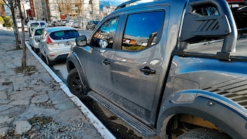 Camioneta gris estacionada en la calle con su rueda delantera contra el cordón de la vereda. Detrás, dos coches blancos y más vehículos. Acera de baldosas irregulares
