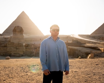 Un hombre con camisa azul claro y gafas de sol posa frente a la Gran Esfinge y las pirámides de Giza con un sol brillante de fondo