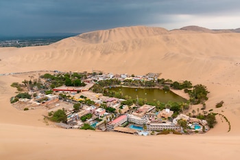 Vista desde lo alto de una laguna verde rodeada por edificios, palmeras y extensas dunas de arena
