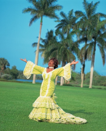 Celia Cruz con los brazos extendidos y un traje verde claro con volantes sobre el césped, rodeada de palmeras bajo un cielo despejado