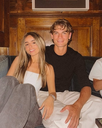 Emma Ramírez y Nicolás Paz posan sonrientes sentados en un sofá gris. Ella viste top blanco y pantalones grises; él, camiseta negra y pantalones claros. Fondo de madera