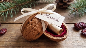 Cáscara de nuez abierta con terciopelo rojo interior, un papel enrollado, una cinta blanca con lazo, una etiqueta con la palabra Mamá y ramas de pino.