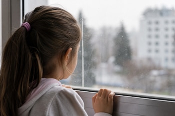 Vista desde atrás de una niña apoyada sobre el marco de una ventana mientras mira hacia el exterior a través del vidrio.
