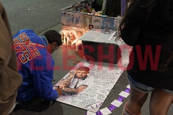 Un hombre escribe sobre un cartel con fotografías y dedicatorias en el suelo, junto a un altar con velas y flores en la vía pública
