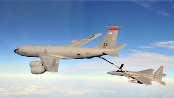 Dos aviones militares grises, un Boeing KC-135 Stratotanker y un caza, vuelan bajo un cielo azul claro mientras el primero reabastece al segundo en el aire