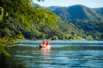 Dos personas con chalecos salvavidas naranjas reman en un kayak naranja en un lago. El fondo muestra montañas verdes cubiertas de árboles y cielo azul