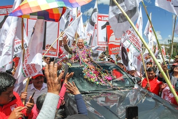 Hombre mayor sonríe y saluda desde el techo de un auto, rodeado de guirnaldas coloridas y numerosas banderas de campaña, en un mitin político con gente