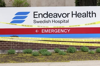 Cartel del Endeavor Health Swedish Hospital y señal de emergencia cubiertos por cinta policial amarilla "POLICE LINE DO NOT CROSS". Abajo, pared de ladrillo y arbustos