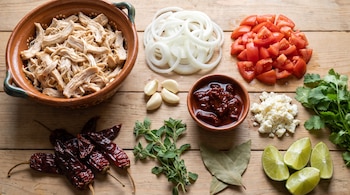 Pollo deshebrado, cebolla en rodajas, jitomate picado, ajos, salsa roja, queso, cilantro, chiles secos, orégano, laurel, limas en tabla de madera.