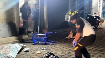 Una persona con uniforme y guantes se agacha en la calle. Hay una silla azul volcada, dos perros, una motocicleta y objetos dispersos