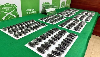 Múltiples paquetes oscuros de droga incautada, envueltos individualmente, se exhiben sobre hojas blancas en una mesa verde con logos de Carabineros de Chile