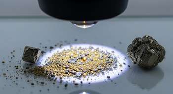 Instrumento negro ilumina con haz de luz un montículo de polvo mineral dorado y gris en una superficie. A los lados, dos fragmentos de pirita.