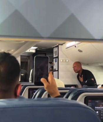 Un pasajero en primer plano graba con su teléfono a un hombre calvo con camiseta negra en un pasillo de avión. Otros pasajeros y asientos de Delta visibles