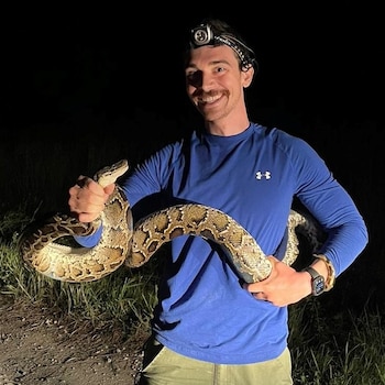 Un hombre con camisa azul y linterna en la cabeza sostiene una serpiente pitón moteada en un camino nocturno