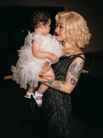 Mujer rubia con vestido oscuro y tatuajes en los brazos sostiene a un bebé que viste un vestido claro con volantes y sandalias blancas. Ambos se miran