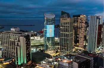 Vista aérea nocturna de una ciudad con rascacielos iluminados, un puerto, barcos y un edificio con letrero Sheraton