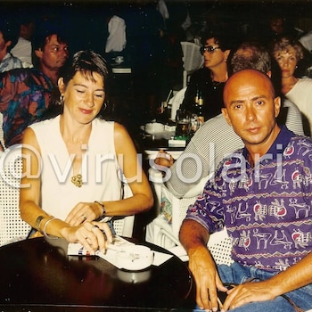 Indio Solari y Virginia sentados en una mesa. Él mira a cámara con camisa estampada púrpura, ella con top blanco y ojos bajos. Fondo borroso