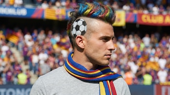 Primer plano de perfil de un hombre con un mohawk multicolor (azul, amarillo, rojo) y un diseño de balón de fútbol pintado en el lateral de su cabeza rapada, llevando una bufanda azul, amarilla y roja.