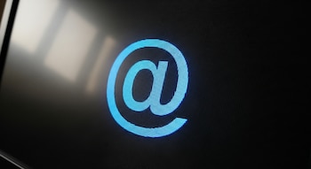 Un primer plano del símbolo arroba azul brillante en una pantalla de computadora con una textura de píxeles visible sobre un fondo oscuro.
