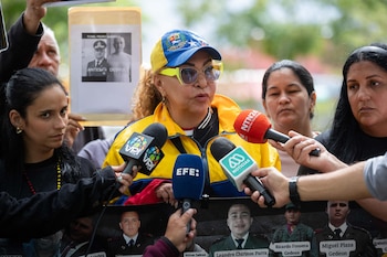Primer plano de mujer con gorra de Venezuela y gafas amarillas hablando a varios micrófonos. Al fondo, personas con carteles y una pancarta con fotos de hombres