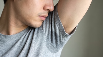 Plano de la parte inferior del rostro de un hombre que acerca la nariz a la axila de su camiseta gris manchada de sudor.