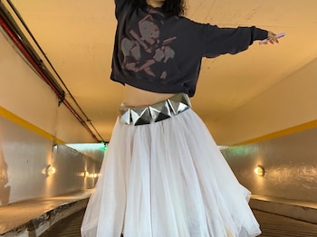 Persona con sudadera oscura, falda de tul blanca y cinturón de tachuelas. Sostiene un teléfono rosa. El fondo es un túnel iluminado