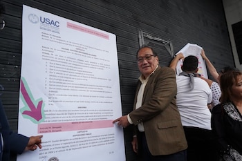 Un hombre con gafas y traje apunta a un gran documento oficial con el logo de USAC y texto sobre leyes electorales. Hay otras personas y una pared metálica