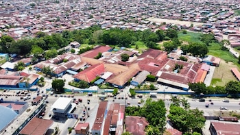 Vista aérea de un complejo de edificios con techos rojizos, rodeado de árboles, una avenida con vehículos y viviendas en el fondo