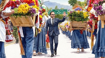 Ilustración de Abelardo de la Espriella saludando en el Desfile de Silleteros con personas llevando flores, un letrero de 'Terminal del Sur' y montañas.