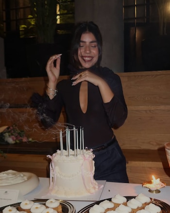 Cami Mayan sonríe sosteniendo un ramo de flores envuelto en papel blanco, vestida con top oscuro, jeans y cinturón dorado, frente a unas escaleras de madera