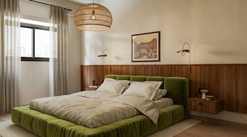 Dormitorio moderno con una cama grande tapizada en verde, ropa de cama clara, cabecero de madera, mesitas de noche y una lámpara colgante de rafia. Ventana lateral con cortinas.