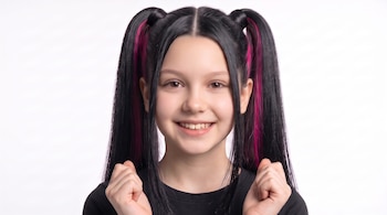 Retrato de una niña sonriente vestida de negro, con coletas altas, mechones fucsias y los puños cerrados frente a un fondo blanco.