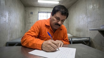 Hombre con bigote, viste mono naranja, escribe una carta con pluma azul sobre una mesa en una celda de concreto.