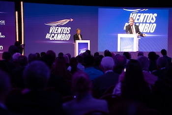 Un hombre de traje habla desde un podio frente a una pantalla iluminada con el texto Vientos de Cambio y un público en penumbra