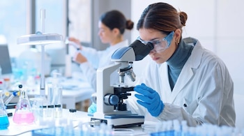 Dos mujeres en batas de laboratorio blancas y guantes, una mirando a través de un microscopio con gafas de seguridad, y la otra trabajando al fondo.