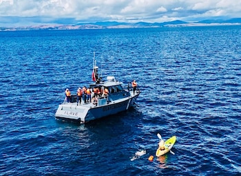 Vista aérea de un nadador junto a un kayakista en un lago azul, con una embarcación de Guardacostas y montañas lejanas bajo un cielo nublado