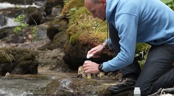 Hombre arrodillado en un arroyo rocoso con musgo, sujetando un frasco de prueba blanco y una botella pequeña con tapa amarilla cerca del agua