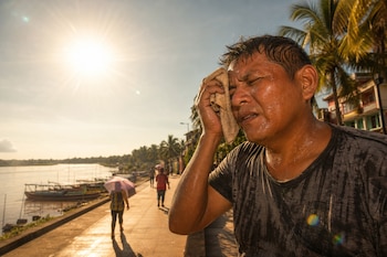 Un hombre sudoroso se seca la frente con un paño bajo un sol intenso junto a un malecón fluvial con palmeras y embarcaciones.