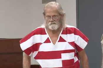 Hombre de edad avanzada con cabello largo y barba blancos, gafas redondas, viste un uniforme carcelario de rayas rojas y blancas, en un recinto interior