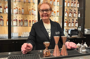 Judy Hollis Jones, con gafas, sonríe detrás de un mostrador de bar con medidores de cócteles de cobre. Detrás hay estantes con botellas de bourbon