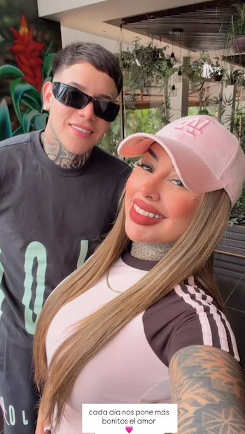 Primer plano de una pareja joven y sonriente con tatuajes. Él usa gafas de sol y camiseta negra; ella, gorra rosa, camiseta bicolor y collar dorado
