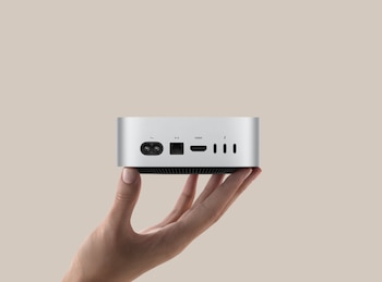 Una mano sostiene la parte trasera de un Apple Mac mini plateado, mostrando los puertos de alimentación, Ethernet, HDMI y Thunderbolt/USB-C sobre un fondo beige