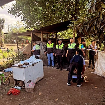 Agentes con uniforme y chalecos reflectantes permanecen de pie junto a una pequeña estructura y un congelador en un terreno arbolado