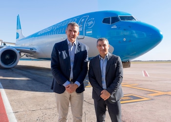 Dos hombres de negocios sonrientes posan de pie frente a la parte frontal de un avión Boeing 737 azul y blanco con el logo de Aerolíneas Argentinas en un aeropuerto soleado