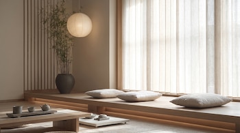 Interior con paredes claras, una plataforma de madera, tres cojines, una mesa baja, un servicio de té, una lámpara de papel, una planta en maceta y persianas verticales.