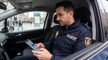 Hombre con uniforme de Policía Nacional sentado en coche, usa teléfono móvil con chatbot; Plaza de España y peatones visibles al fondo.