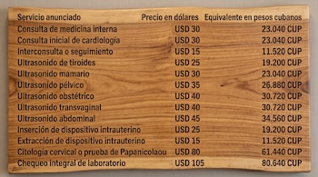 Tabla sobre fondo de madera con tres columnas que listan servicios médicos y sus precios en dólares y pesos cubanos.