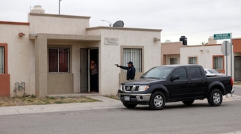 Hombre de chaqueta oscura apunta un arma a una mujer en la puerta de una casa, junto a una camioneta pick up negra. Se observan letreros de calles.